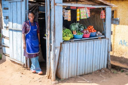 Eine junge Frau mit Schürze steht neben einer kleinen, blaugestrichenen Bretterbude, auf deren Auslage Obst und Gemüse liegen.