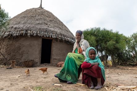 Eine Frau und ein kleines Mädchen sitzen auf dem Platz vor einer strohgedeckten, runden Lehmhütte. Einige Hühner laufen um sie herum.