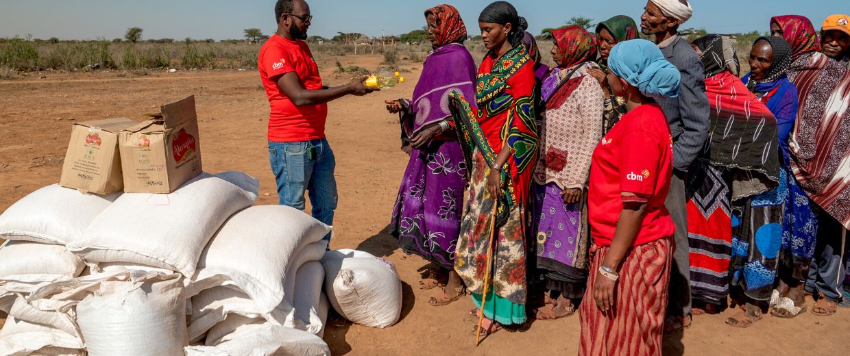 Buntgekleidete Menschen stehen Schlange vor einem Stapel aus weißen Säcken. Ein Mann im roten CBM-T-Shirt überreicht der ersten Frau in der Schlange eine Speiseölflasche.