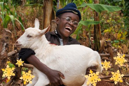 Ein lachender Junge mit Nickelbrille und Mütze hält eine Ziege im Arm.