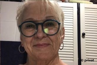 Porträt einer Frau mit weißer Kurzhaarfrisur und Brille vor einem weißen Schrank mit Lamellentür.