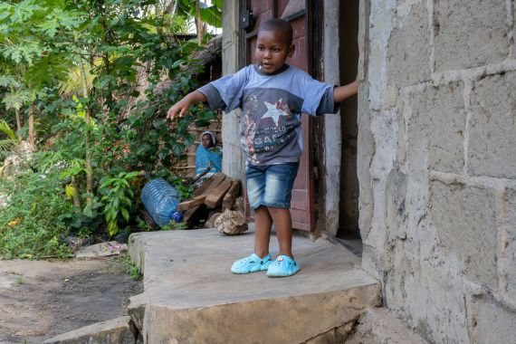 Ein kleiner Junge steht auf einer Betonschwelle vor einem Hauseingang und tastet sich mit einer Hand an der Wand entlang.