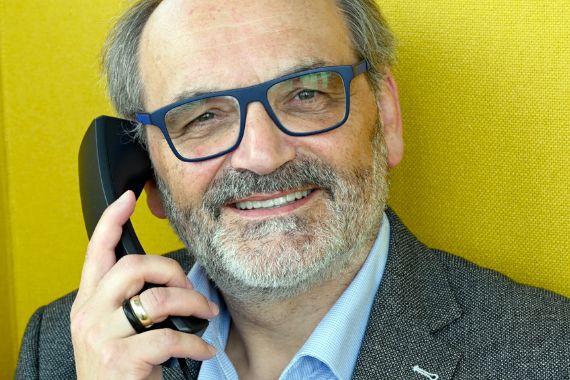 Ein etwa 60-jähriger lächelnder Mann mit einer blauen Brille hält sich einen Telefonhörer ans Ohr.