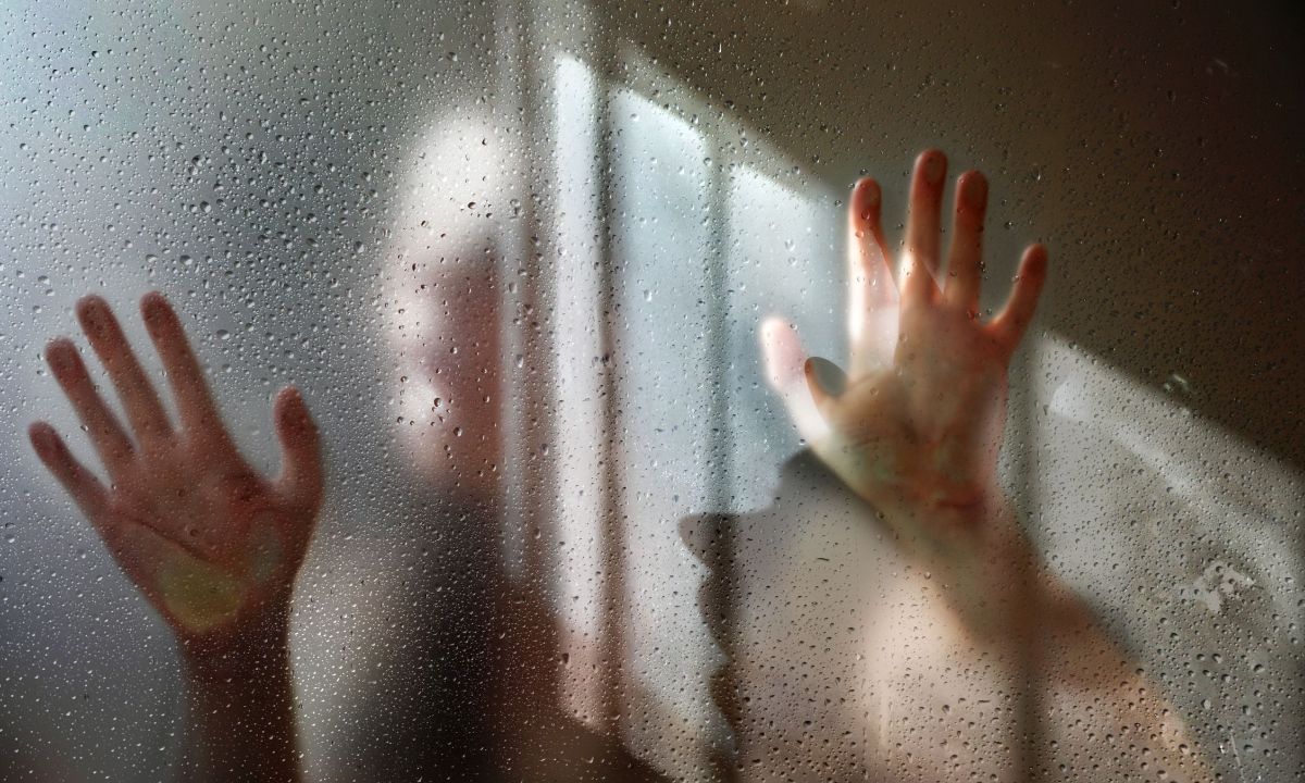 Zwei Hände auf einer nassen Glasscheibe, dahinter die Silhouette einer Frau. Auf der Scheibe spiegelt sich ein Fenster. Ein Lichtschein wirft den Schatten der Frau auf die Scheibe.