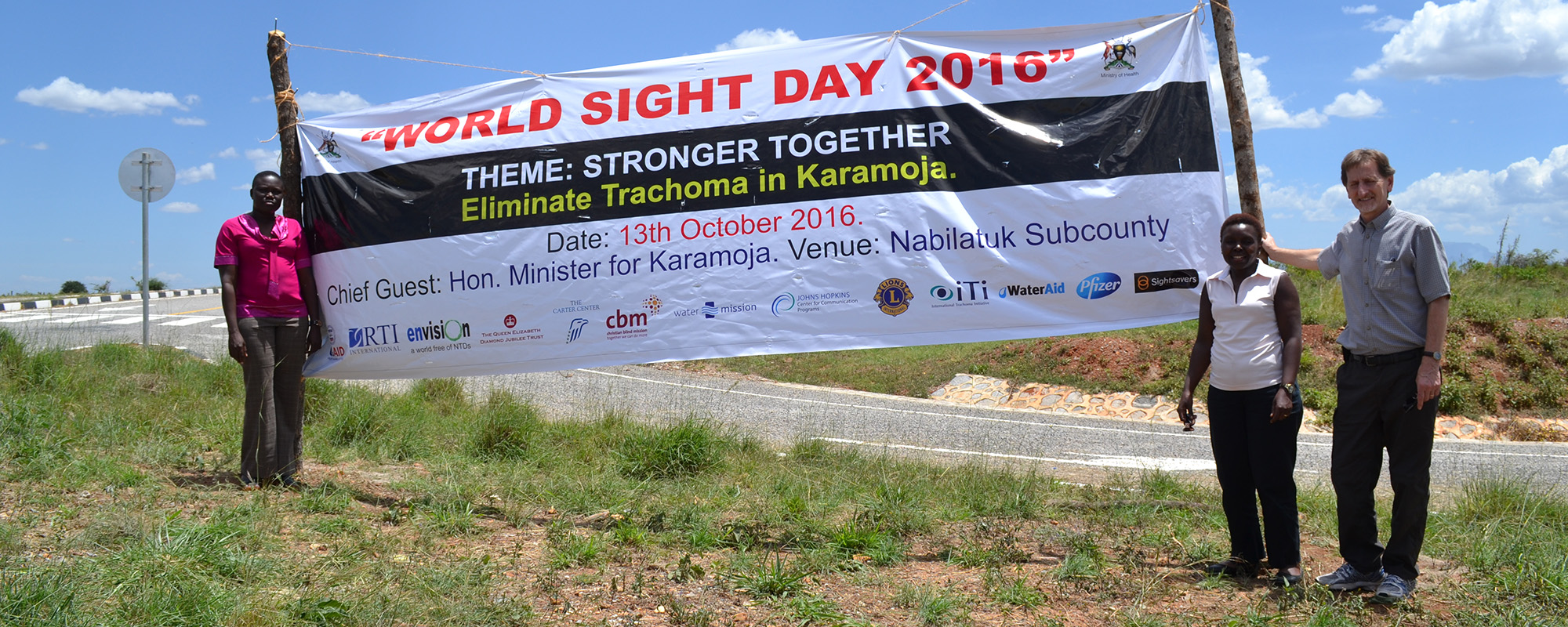 Drei Menschen stehen neben einem Plakat, auf dem über einen medizinischen Außeneinsatz für Augenpatienten in Karamoja/Uganda am Weltsehtag 2016 informiert wird.